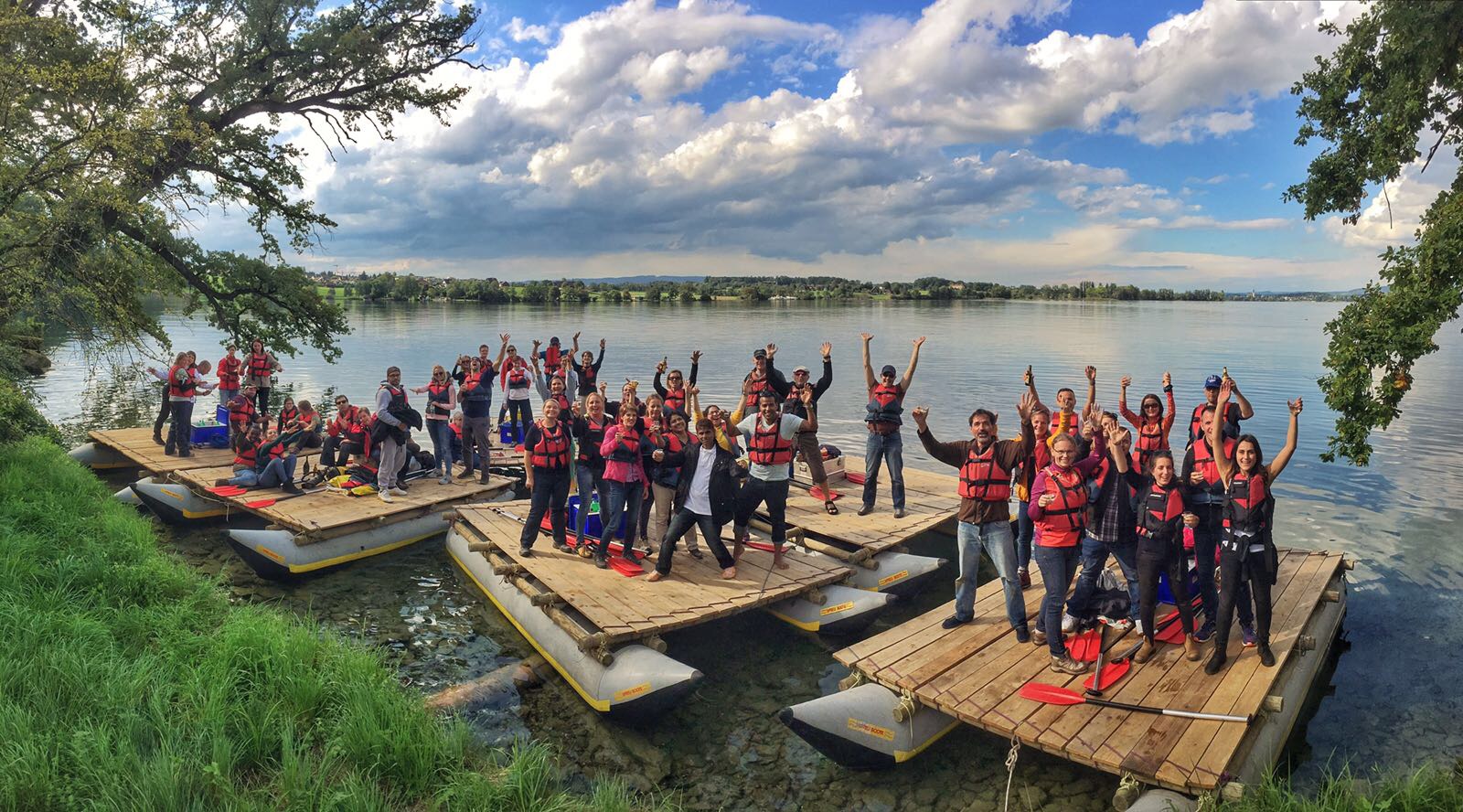 Am Ufer sind 6 Flosse mit jeweils einer Gruppe von Leuten drauf, alle schauen in die Kamera und halten die Hände hoch