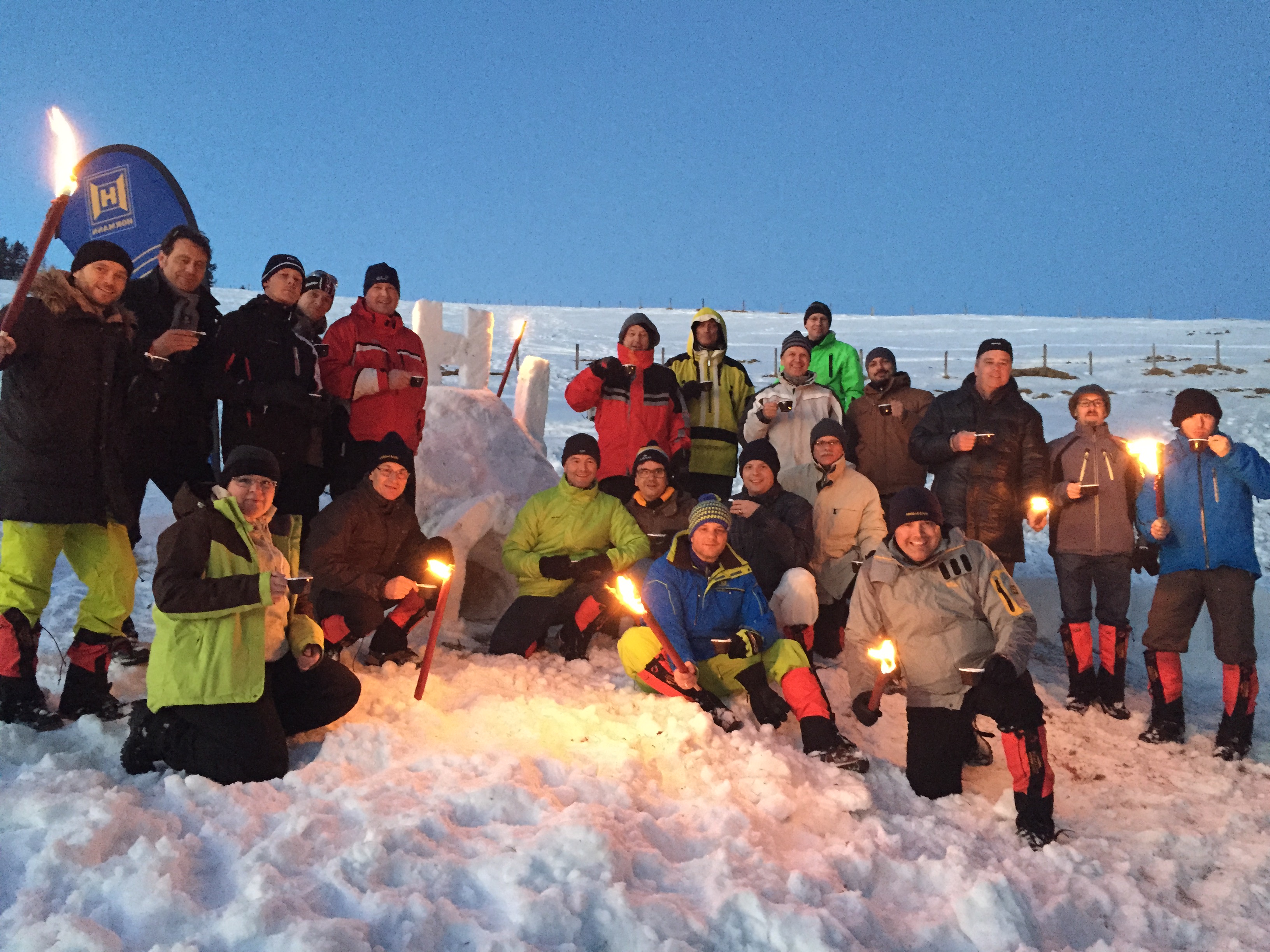 Ein Gruppenfoto im Schnee, es ist nach Sonnenuntergang und die Leute halten zum Teil Fackeln und einen Becher mit heissem Getränk