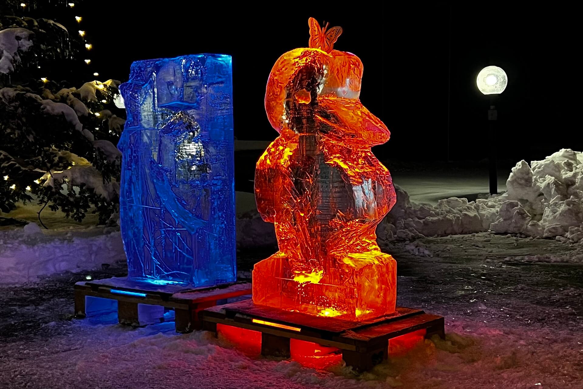 2 Eisskulpturen bei Nacht, die rechte ist ein Pinguin welche rot beleuchtet ist, die linke ist eine Sternschnuppe, die blau beleuchtet ist.