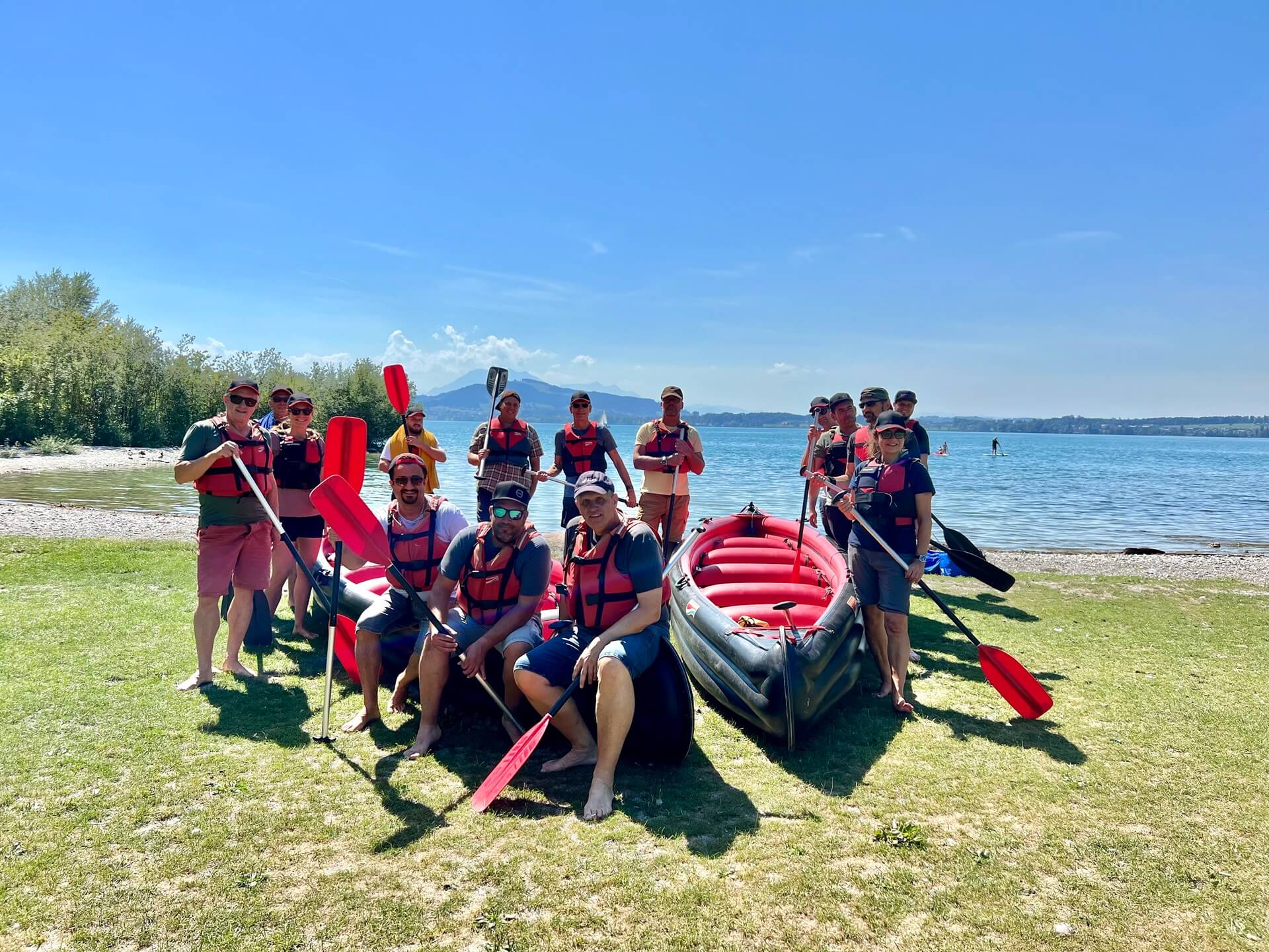 Gruppenfoto am Ufer mit 2 Grosskanadier im Rasen, auf dem linken Kanu sitzen 3 Leute, weitere Leute (12) stehen um die zwei Kanus herum, alle halten ein Paddel in der Hand