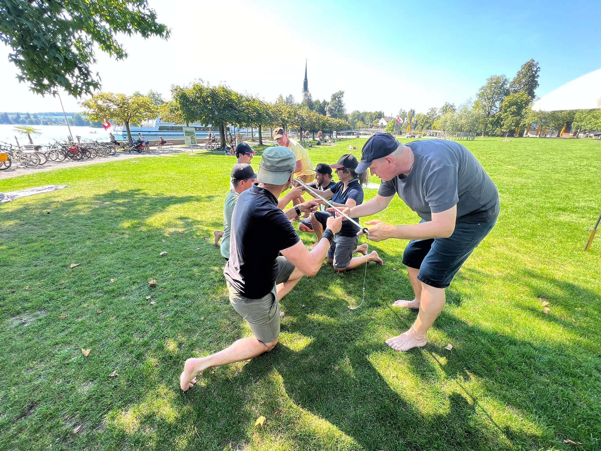 Eine Gruppe im Park macht eine Gruppenübung, sie stehen parallel zu einander und balancieren in der Mitte einen Stab.