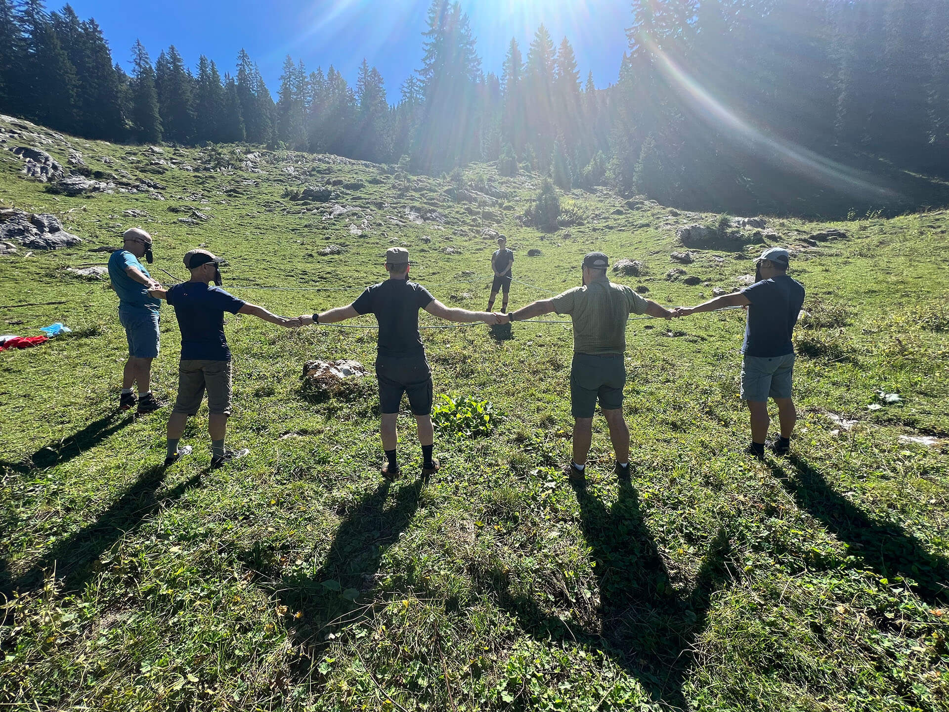Eine Gruppe steht am Waldrand im Kreis, sie halten ein Seil und ihre Augen sind verbunden.