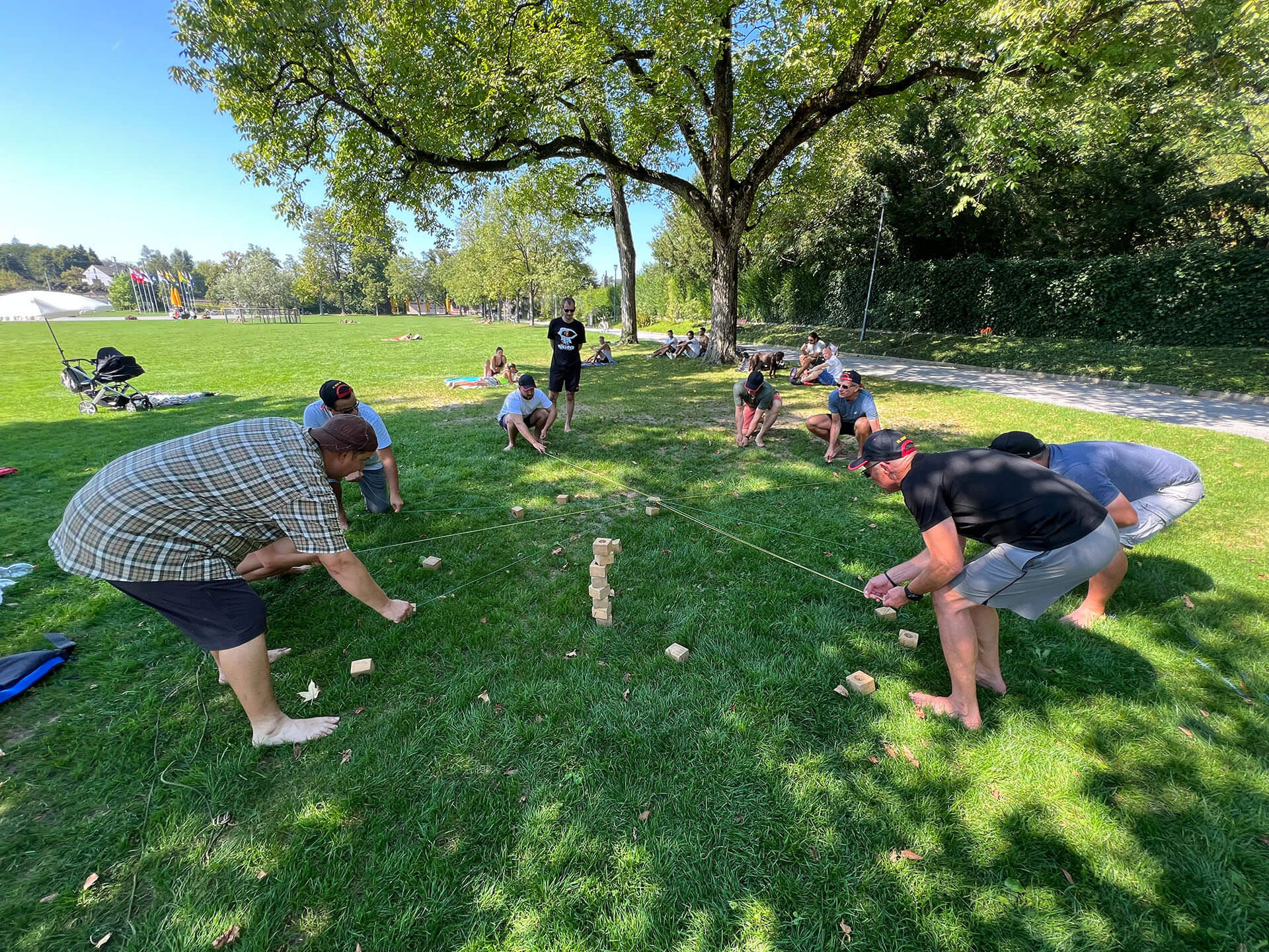 Eine Gruppe im Park macht eine Gruppenübung, in der Mitte ist ein Turm aus Holzklötzchen, die Teilnehmer halten eine Schnur welche in der Mitte zusammengemacht ist und versuchen das nächste Klötzchen auf den Turm zu legen.