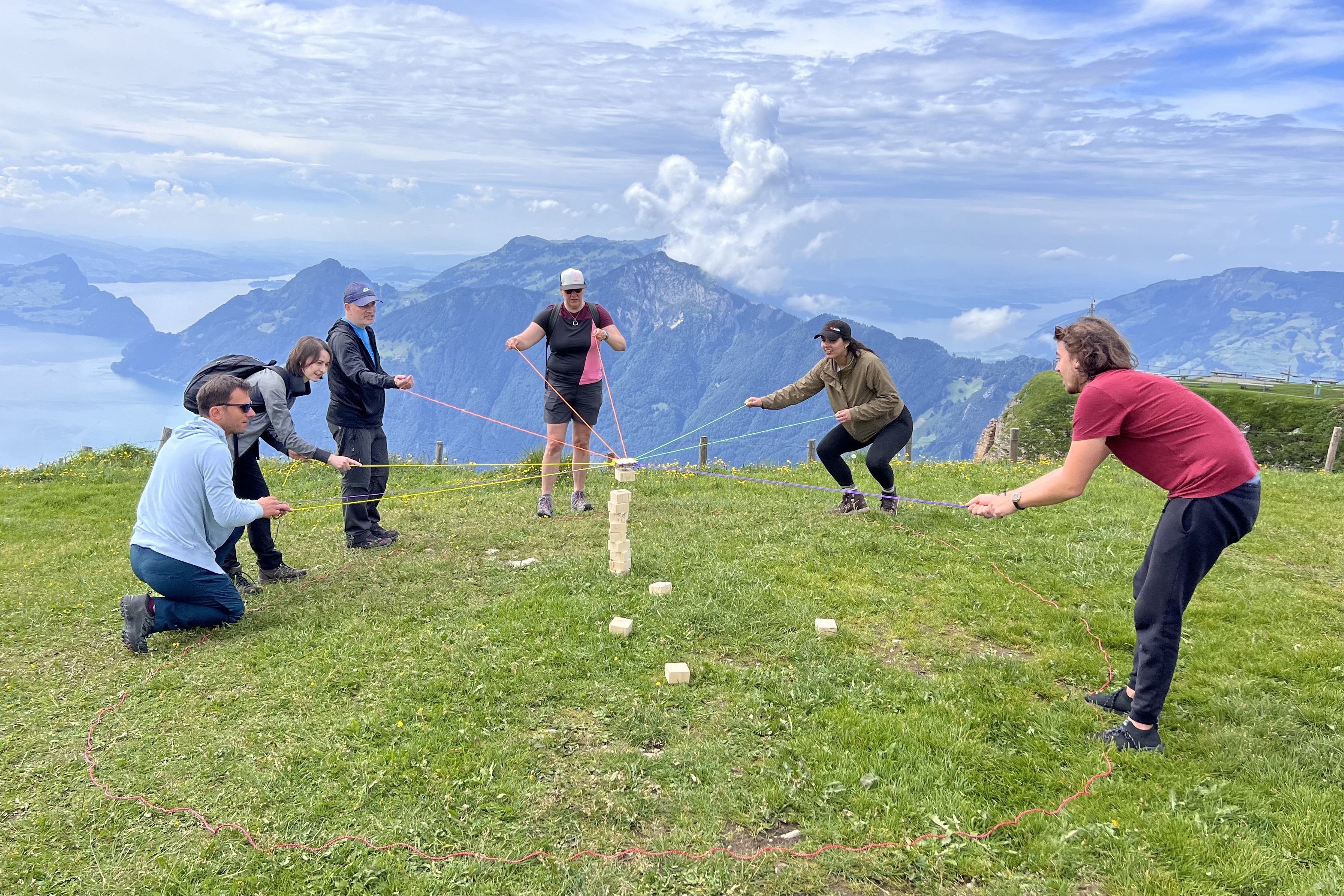 Eine Gruppe auf einer Wiese macht eine Gruppenübung, in der Mitte ist ein Turm aus Holzklötzchen, die Teilnehmer halten eine Schnur welche in der Mitte zusammengemacht ist und versuchen das nächste Klötzchen auf den Turm zu legen.