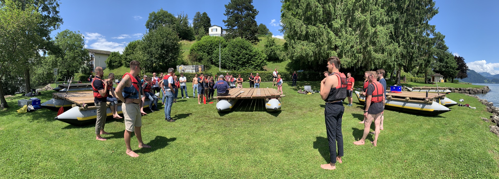 Auf einer Wiese am See stehen verteilt 5 Flosse,  um das mittlere Floss stehen Leute die Instruktionen erhalten