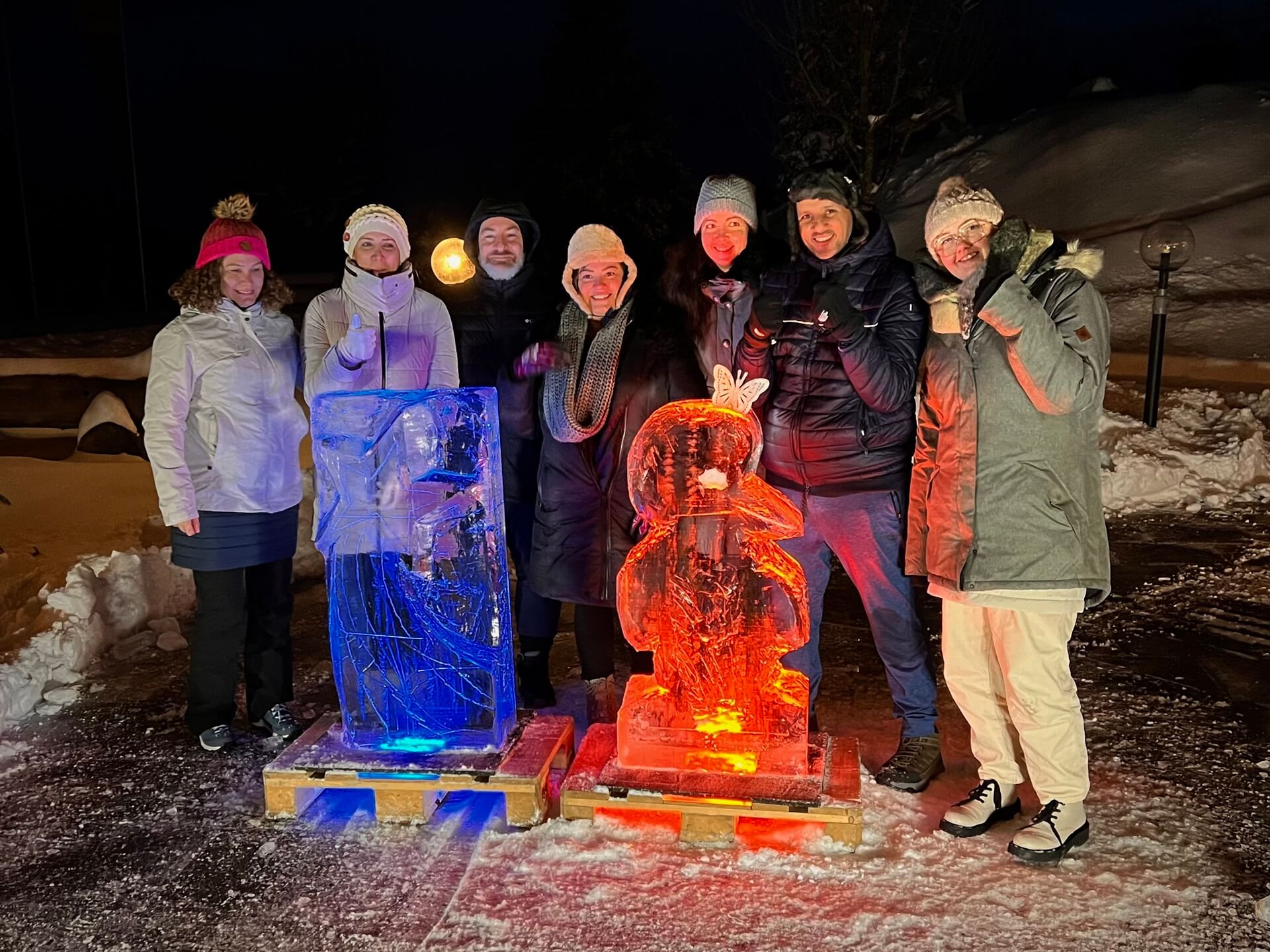 Sieben Leute stehen hinter 2 Eisskulpturen, es ist Nacht und die Skulpturen sind farbig beleuchtet, eine blau und die andere rot