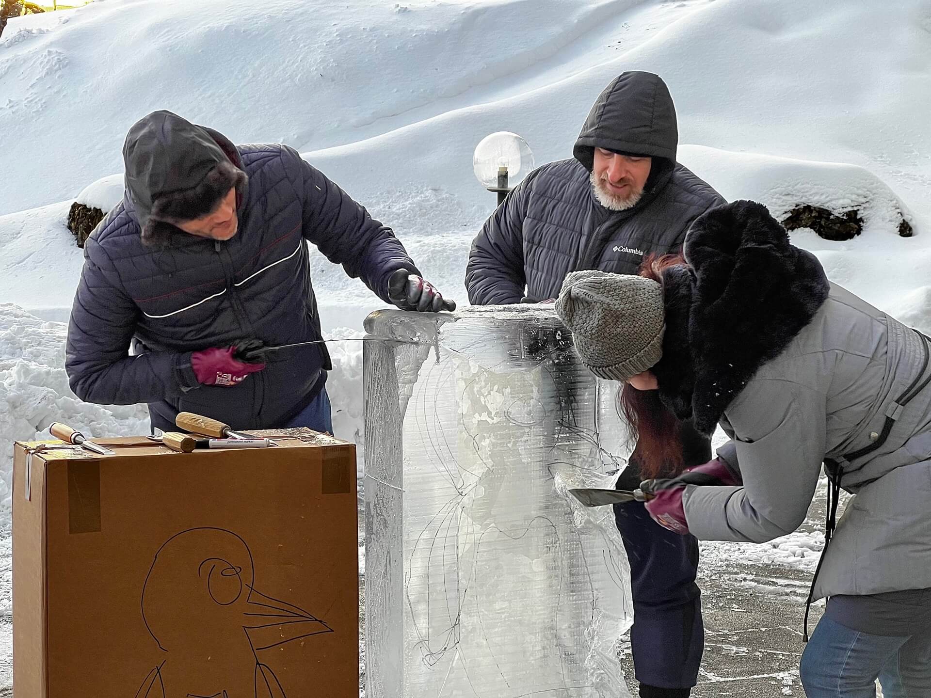 Drei Leute arbeiten an einer Eisskulptur, eine Person hält eine Säge und eine andere einen Spachtel