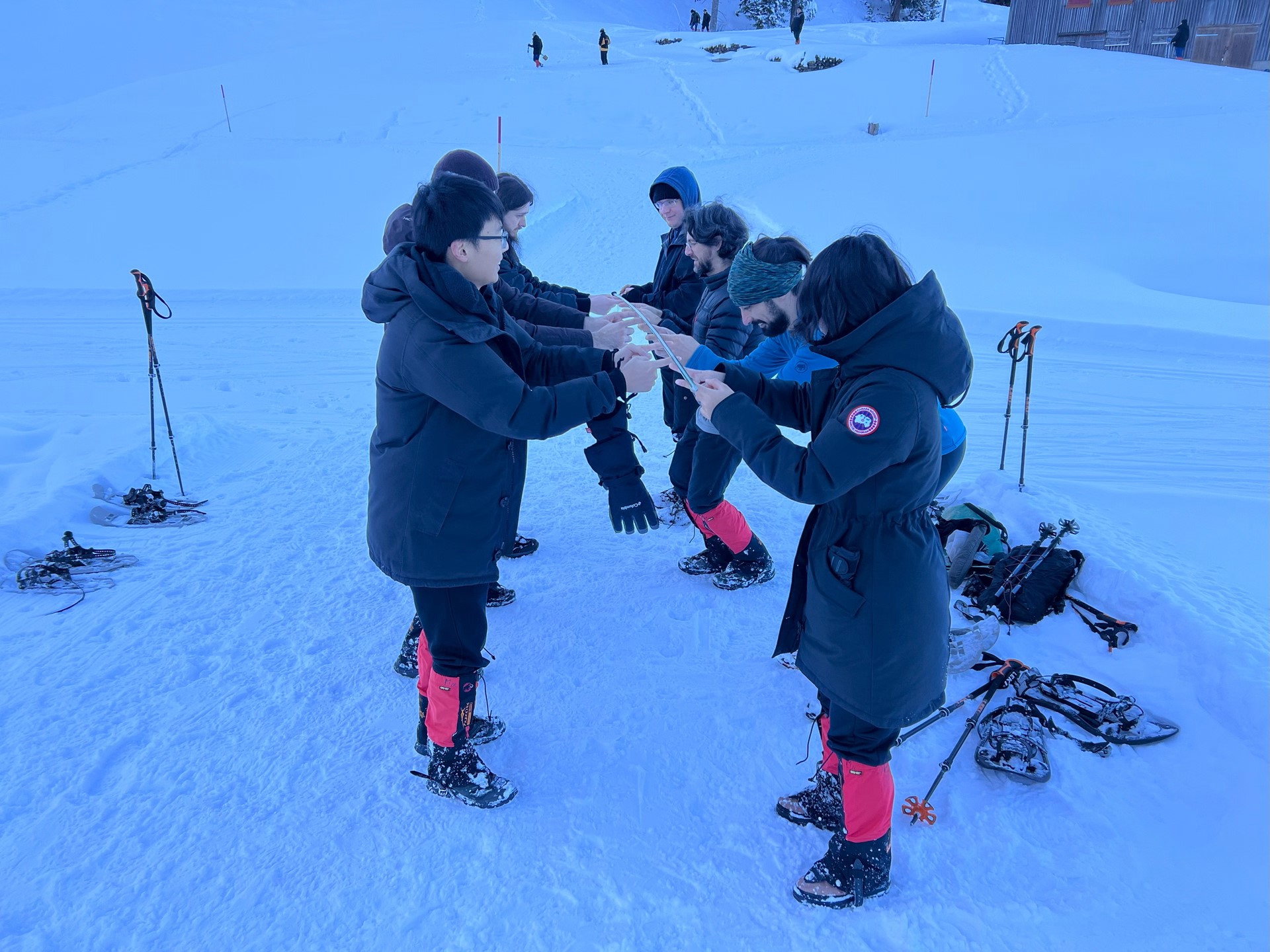 Sieben Leute stehen vis à vis zu einander im Schnee und balancieren zwischen ihnen einen Stab auf den Finger